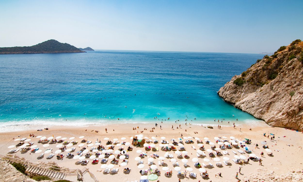Mala-Strand in der Türkei: Eine Bucht mit türkisblauem Meer, felsiger Steilküste und hellem Sandstrand voller weißer Sonnenschirme.