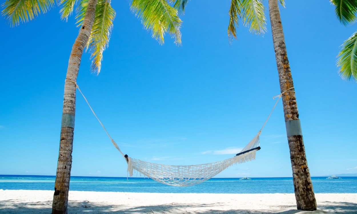 Netzhängematte zwischen zwei Palmen an einem weißen Sandstrand vor türkisblauem Meer und klarem, blauem Himmel.