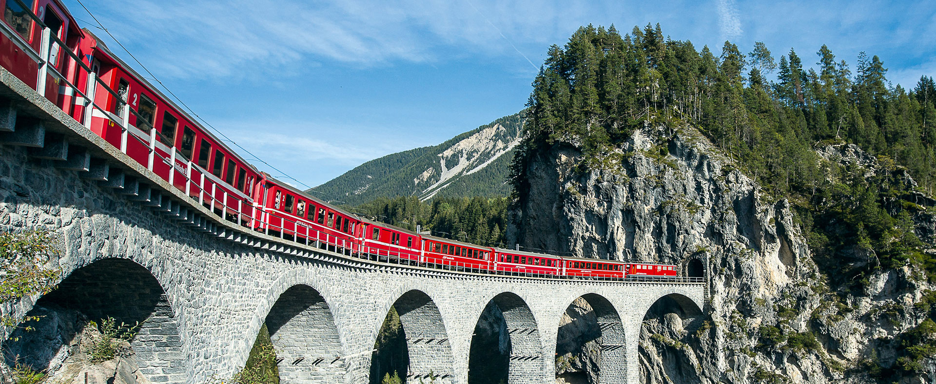 Ein roter Zug der Rhätischen Bahn fährt am Ufer des Flusses durch die beeindruckende Ruinaulta Schlucht in Graubünden. Die Szene wird von hohen, steilen Felswänden in hellen Grau- und Weißtönen dominiert, die sich dramatisch über den Fluss erheben. Einzelne Bäume und grüne Vegetation sind auf den Felsen und am Flussufer zu sehen. Der Himmel ist teilweise blau mit einigen weißen Wolken.